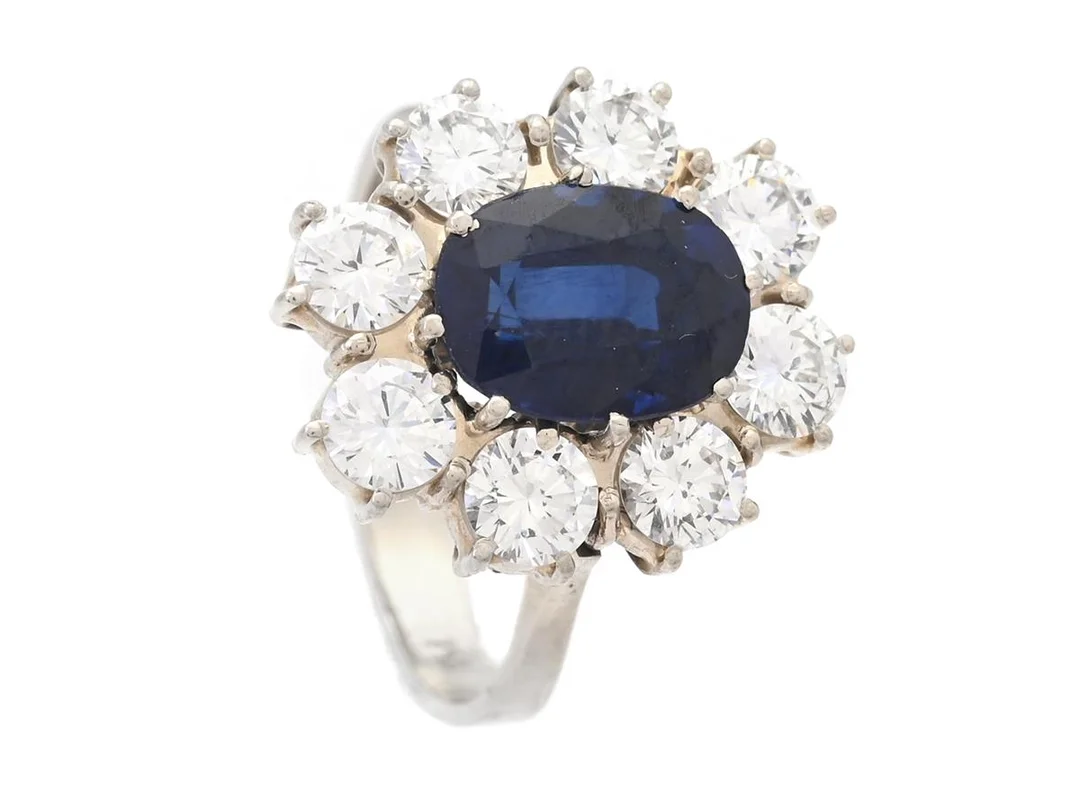 CARMOSÉRING, 18K rodinerat guld, ovalslipad syntetisk blå safir ca 2,50 ct, 8 briljantslipade diamanter ca 1,60 ctv, ca TW/VS, stl 17,25 mm, vikt 5,5 g, safir med nagg och skador, en diamant med nagg vid rondisten. — Smycken/Carmoséring — Kaplans