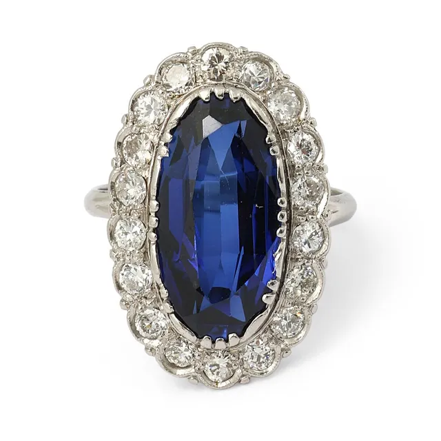 A sapphire and diamond ring set with an oval-cut synthetic sapphire encircled by numerous brilliant-cut diamonds, mounted in platinum. Weight app. 4.5 g. W. 23 mm. Size 51.