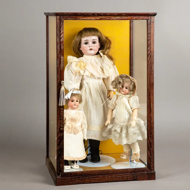 Porzellankopfpuppen (3) in Glasvitrine um 1900.