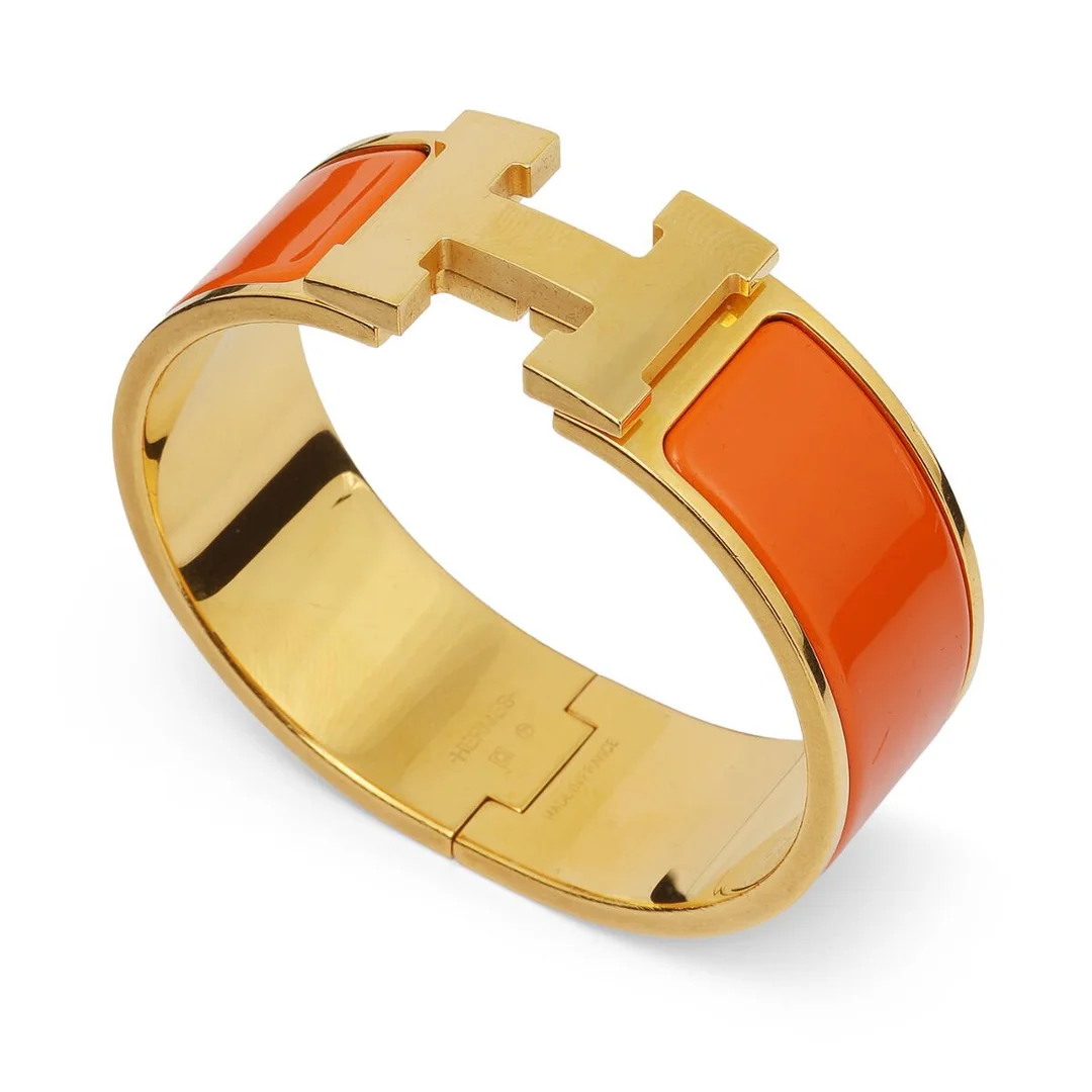 A “Clic Clac H” bangle of gold coloured metal and orange enamel. Weight app. 36 g. W. 12 mm. Inner diam. app. 5.5 cm. — Designer Handbags & Fashion — Bruun Rasmussen