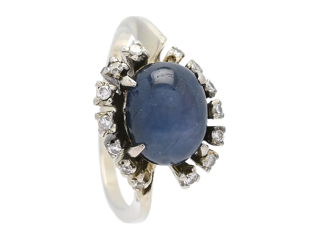 RING, 18K vitguld, cabochonslipad blå safir ca 3,50 ct med svag stjärna, sprickor, 14 briljanter ca 0,14 ctv, 5 stenar skadade, stl 17,75 mm, vikt 5,9 g. — Smycken/Ring — Kaplans