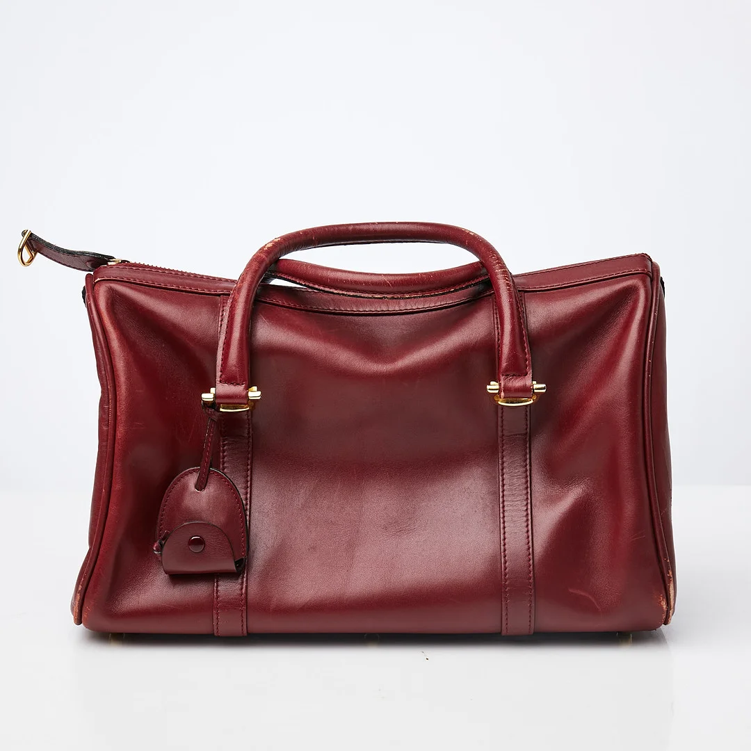 LES MUST DE CARTIER. Handväska, slätt vinrött skinn, dragkedsförsedd, med top handle-handtag, detaljer i gulmetall, en dragkedsförsedd innerficka, märkt med logo samt på plakett "les must des Cartier Rd 1975 Paris". — Vintage & Designer Fashion — Stockholms Auktionsverk Sickla