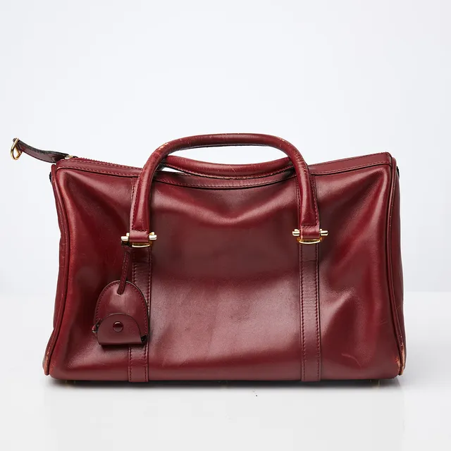 LES MUST DE CARTIER. Handväska, slätt vinrött skinn, dragkedsförsedd, med top handle-handtag, detaljer i gulmetall, en dragkedsförsedd innerficka, märkt med logo samt på plakett "les must des Cartier Rd 1975 Paris".