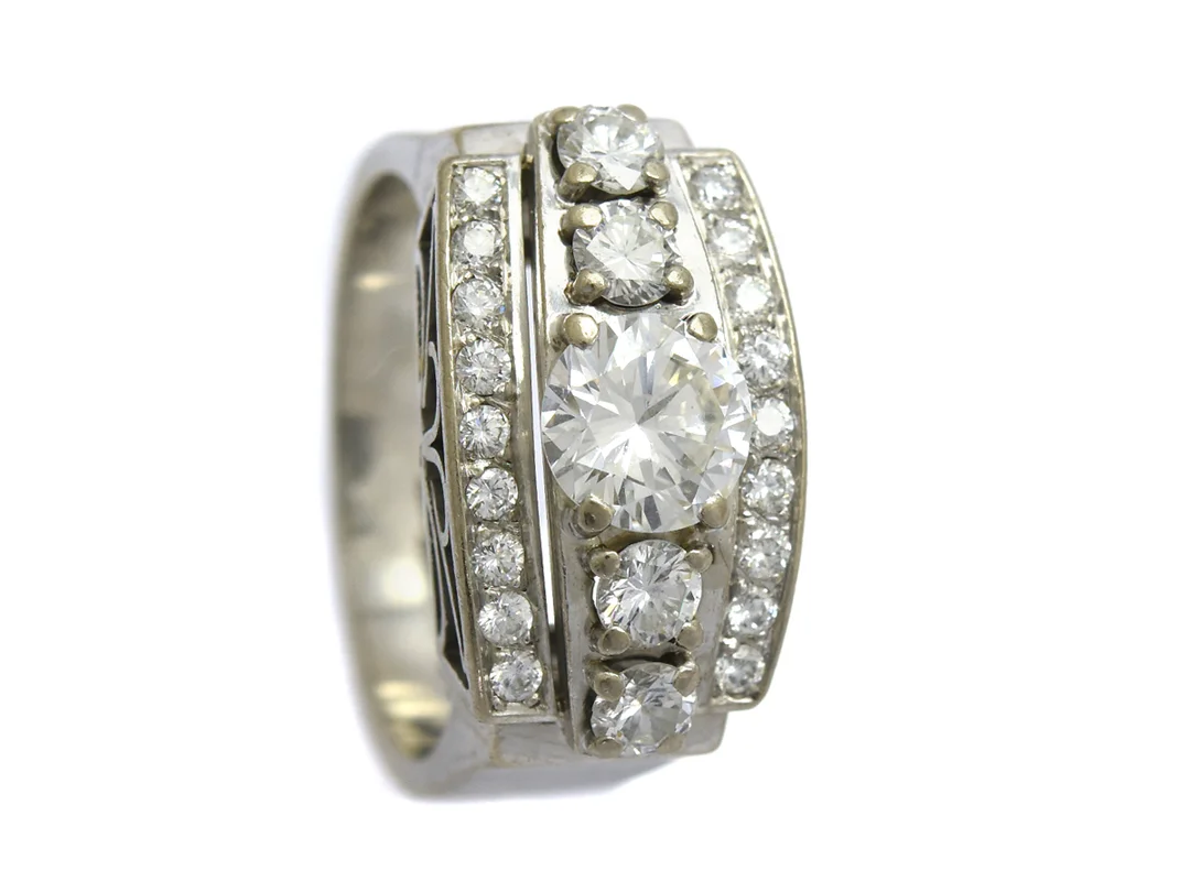 RING, 18K vitguld, briljantslipad diamant ca 0,94 ct, ca H-I(W-TCR/SI (nagg i rondisten) 22 briljantslipade diamanter ca 0,90 ctv, ca TW-W/VS, stl 17,75 mm, Björkman & Magnell, Stockholm 1985, vikt 12,4 g, mittstenen sitter löst i sin fattning, tre av de fyra större sidostenarna är skadade. — Smycken/Ring — Kaplans