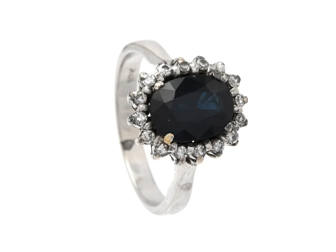 CARMOSÉRING, 18K vitguld, ovalslipad mörkblå safir, 16 åttkantslipade diamanter ca 0,16 ctv, stl 16,25 mm, vikt 4,1 g, safiren med små nagg, slitna klor. — Smycken/Carmoséring — Kaplans