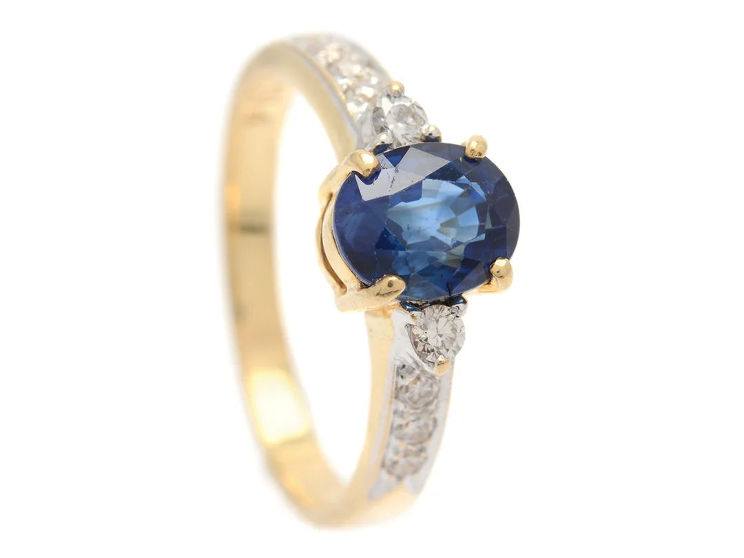 RING, 18K guld, ovalslipad blå safir ca 1,84 ct, 8 briljantslipade diamanter ca 0,20 ctv, ca Cr(J)/SI, stl 17 mm, vikt 3,7 g, safiren har nagg vid rondisten och slitna fasettkanter. — Smycken/Ring — Kaplans