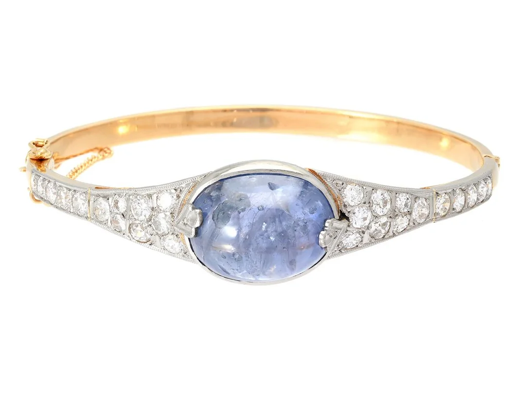 ARMRING, 18K guld/vitguld, cabochonslipad safir ca 25,00 ct, gammal- och briljantslipade diamanter ca 2,00 ctv, ca TW-Cr/VS-P, Stockholm 1950, stl 56x52 mm, vikt 27,0 g, säkerhetskedja, tennlödningar, en del skev, safiren har naturliga håligheter och inneslutningar som når ytan, enstaka diamanter har nagg och skador. — Smycken/Armring — Kaplans