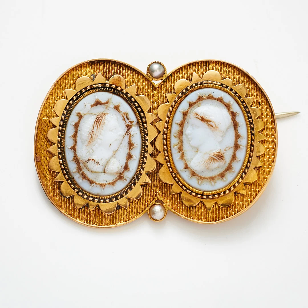 BROSCH, 18 k guld, 2 infattade parställda kaméer av vit glasmassa föreställande klassiska kvinnoprofiler, 2 orientpärlor, 1800-talets andra hälft. — Brooches & Pendants — Stockholms Auktionsverk Magasin 5